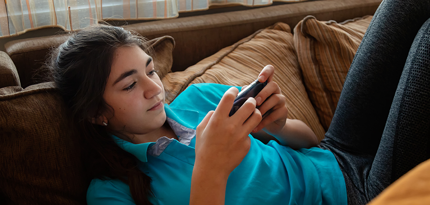 Teen girl lying on brown couch and scrolling her phone, browsing social media