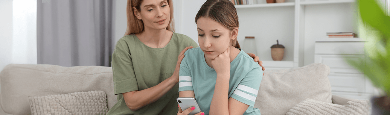 teenage girl on sofa looks sadly at her phone while mother comforts her by holding her shoulders