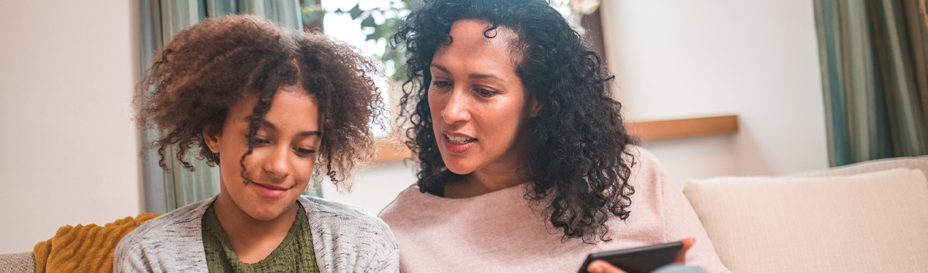 mother looking over teenage daughter's shoulder on couch as she helps to adjust settings on her child's tablet device